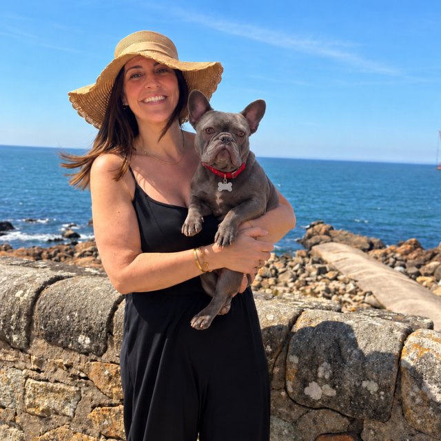 Leighann Huckstep in a sun hat by the ocean, holding her grey French bulldog, Gus.